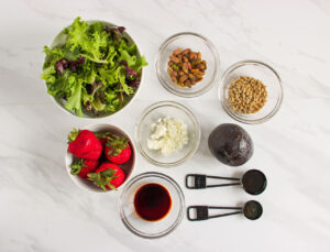 Strawberry Pistachio Goat Cheese Salad ingredients in individual bowls on a white marble surface.