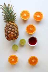 Tequila Sunrise Mocktail recipe ingredients laying on a white surface. A whole pineapple, cut citrus and a white bowl of grenadine.