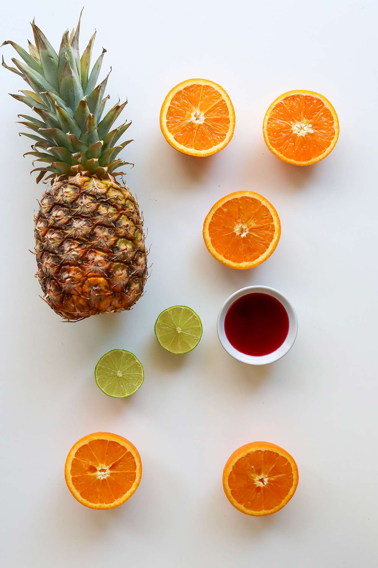 Tequila Sunrise Mocktail recipe ingredients laying on a white surface. A whole pineapple, cut citrus and a white bowl of grenadine.