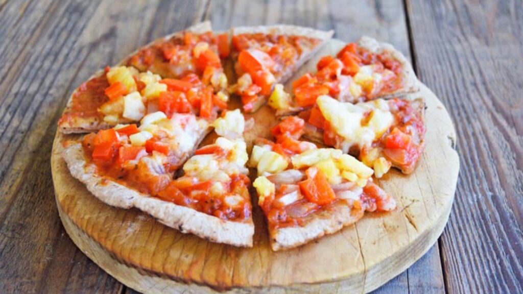 A Hawaiian pita pizza sits on a round cutting board on a wood table.