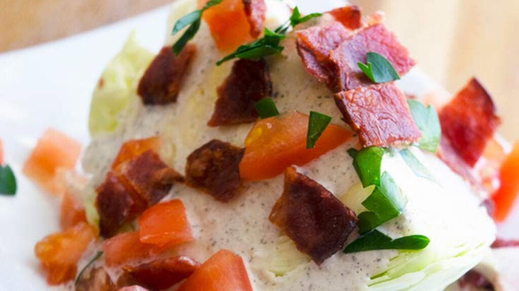 A salad wedge sits on a white plate smothered in bacon bits, chopped tomatoes and ranch dressing.