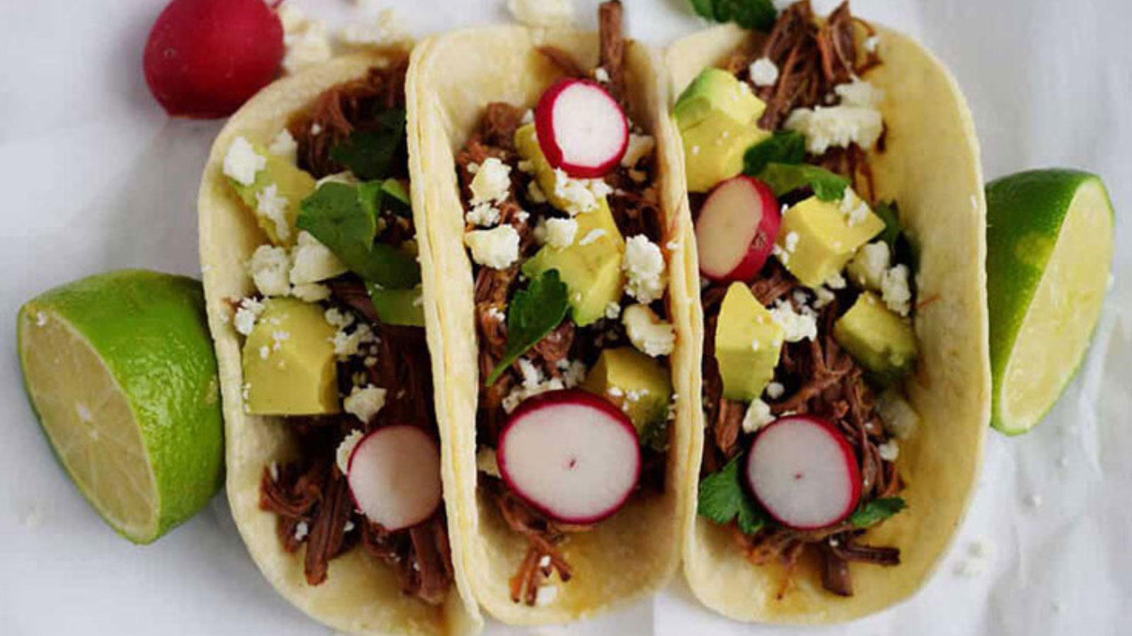Three tacos lined up on a white surface filled with beef brisket and taco toppings.