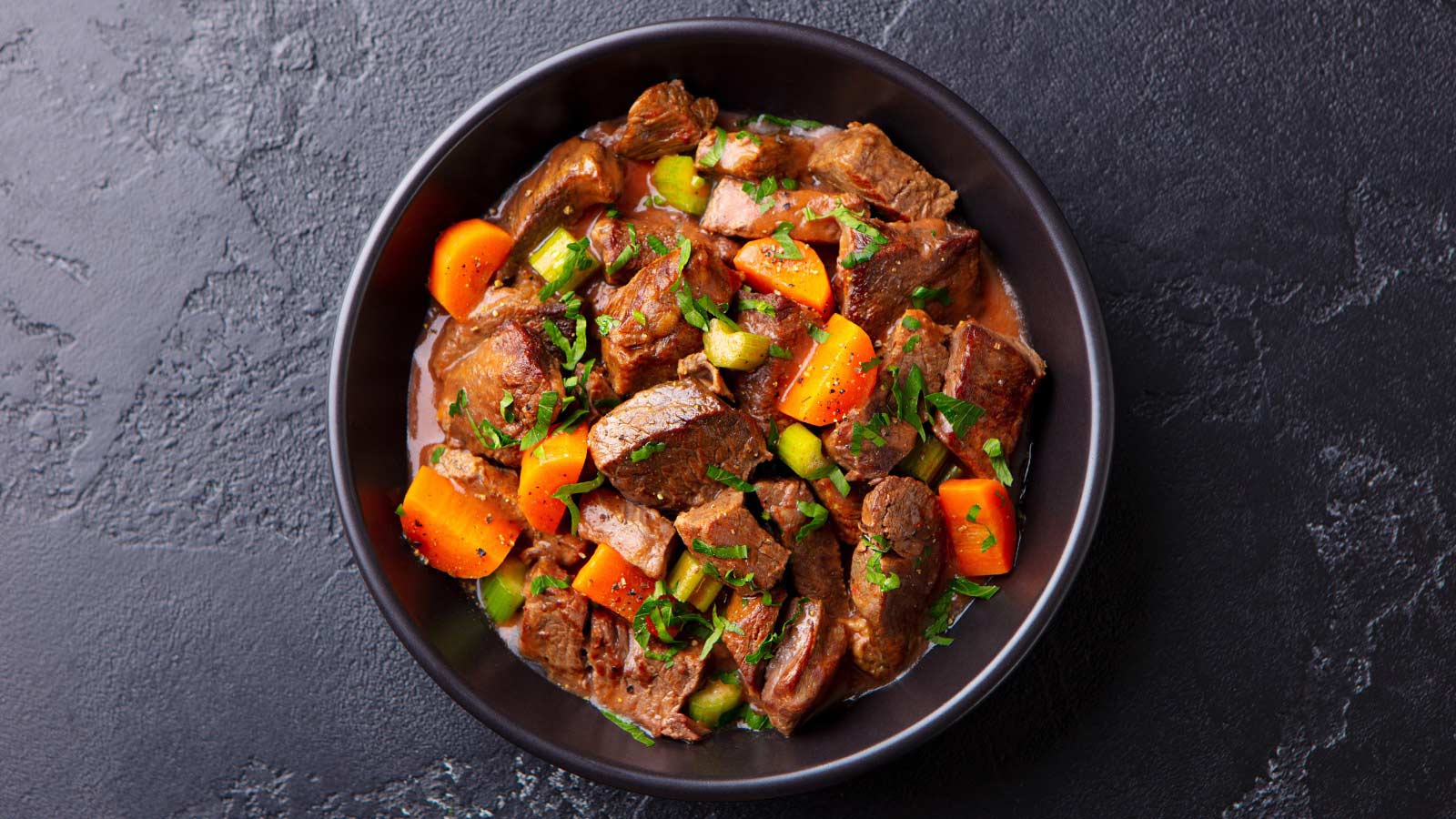 Beef meat and vegetables stew in black bowl. Slate background. Top view.