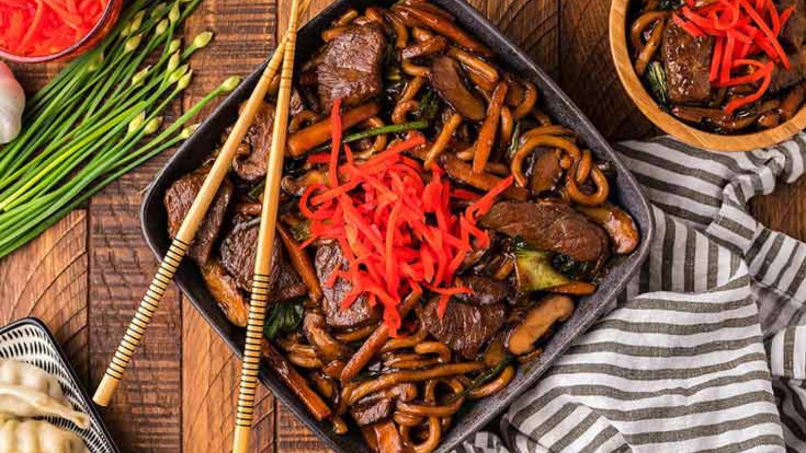 An overhead view of a black, square plate filled with beef yaki udon noodle stir fry topped with shreds of red bell pepper.