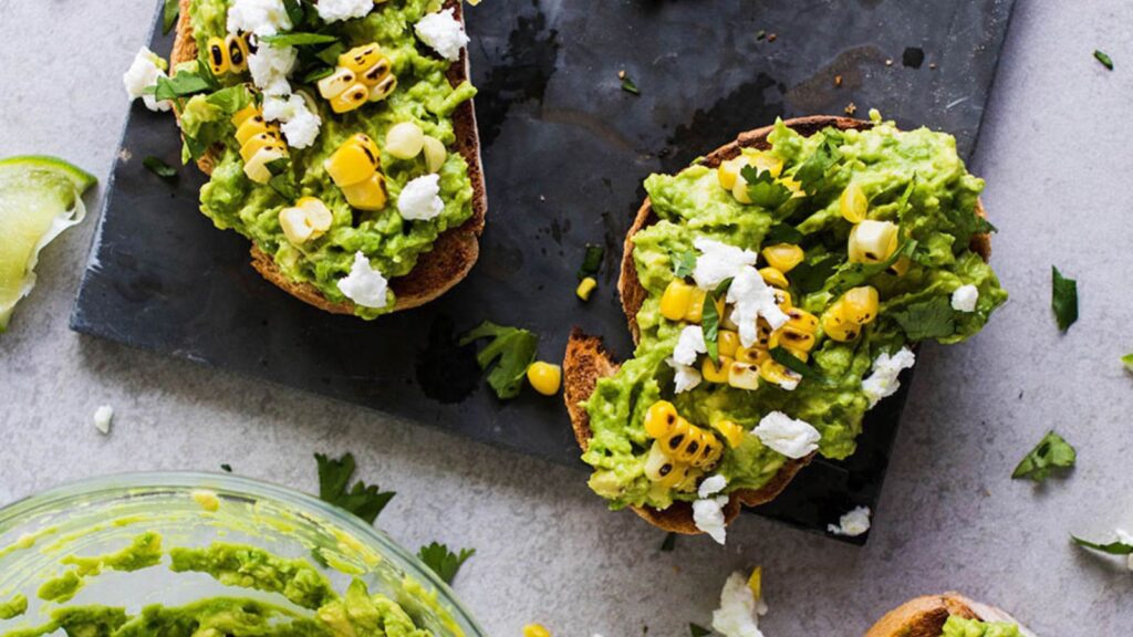 An overhead view of two slices of grilled corn avocado toast on a piece of slate.