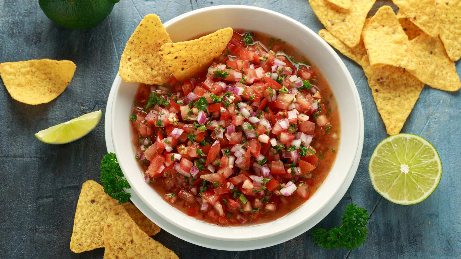 Mexican Tomato Salsa in white bowl with lime, red onion, jalapeno pepper, parsley and tortilla chips.