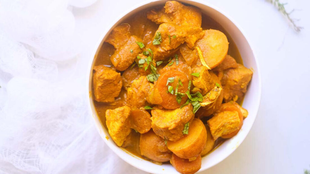 A white bowl holds Jamaican Curry Chicken with large chunks of chicken, potatoes and carrots.