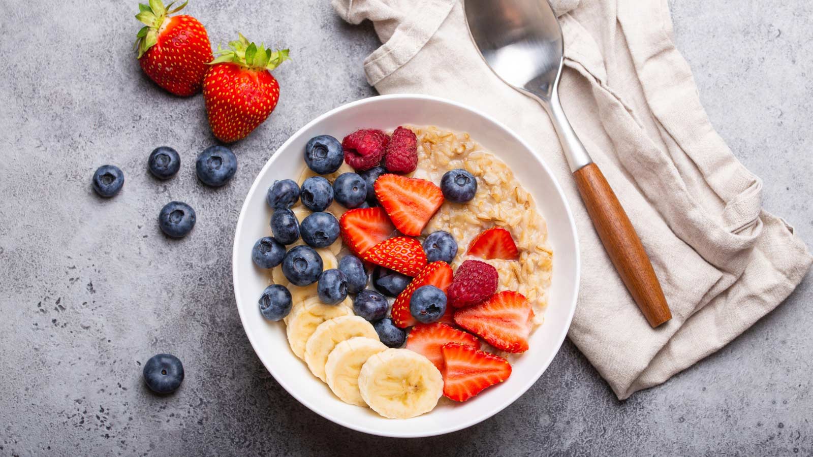 Oatmeal porridge with fruit and berries in bowl with spoon on white wooden background table top view, homemade healthy breakfast cereal with strawberry, banana, blueberry, raspberry