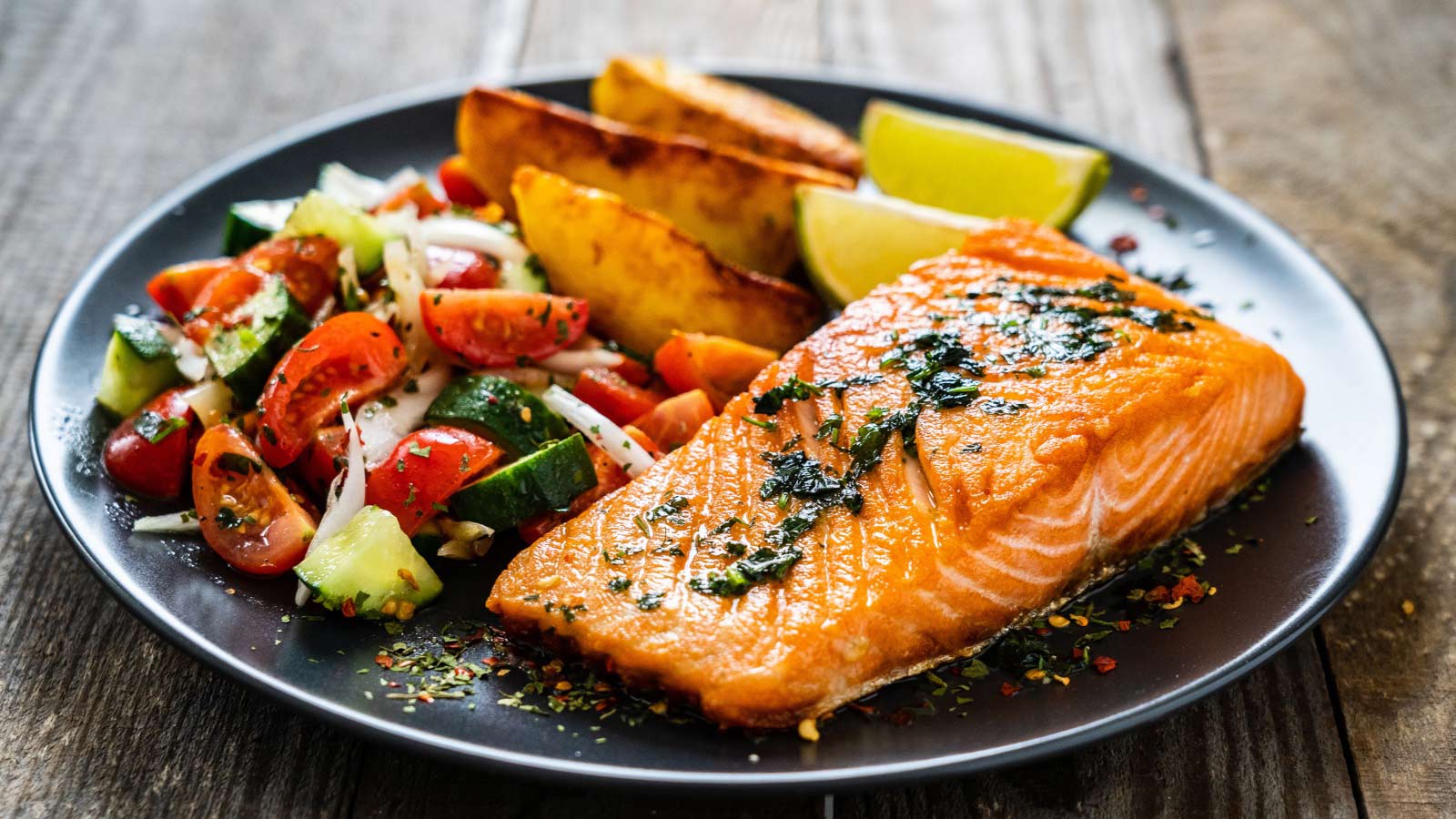 Fried salmon fillet with fried potatoes, lime and vegetable salad served on black plate on wooden table.