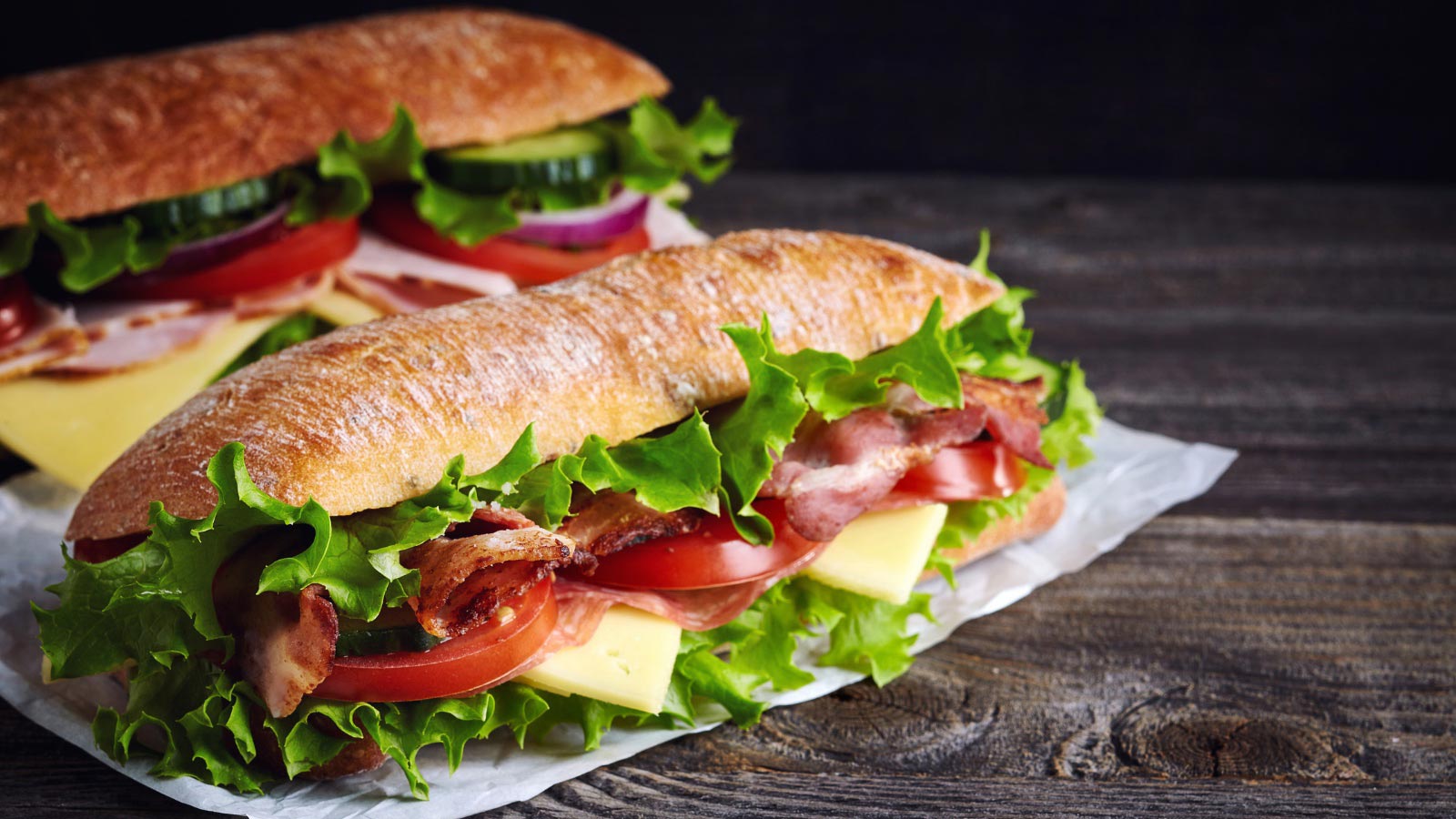 Two fresh submarine sandwiches with ham, cheese, bacon, tomatoes, lettuce, cucumbers and onions on dark wooden background.