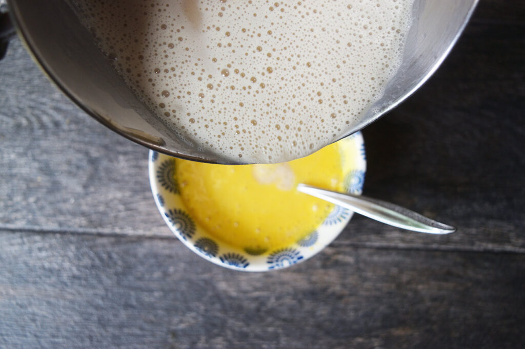 Pouring hot milk into a small bowl of egg yolks to temper them.