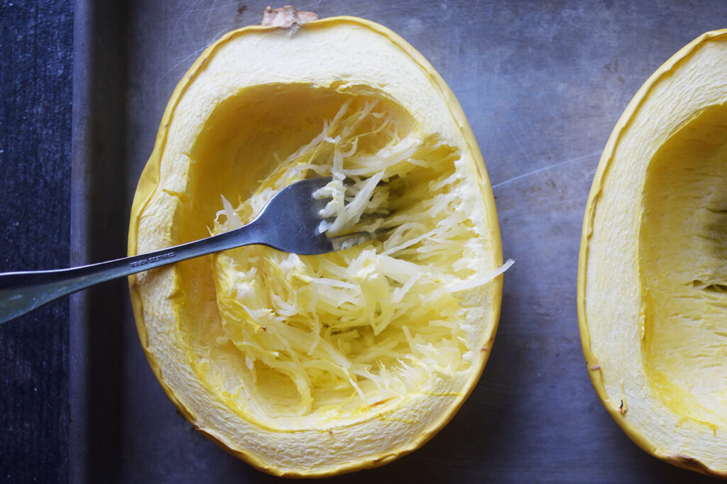 Scraping cooked spaghetti squash crosswise to loosen the flesh.