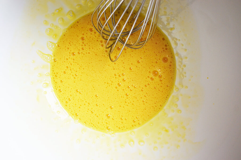 A white mixing bowl with whisked eggs and a whisk sitting in it.