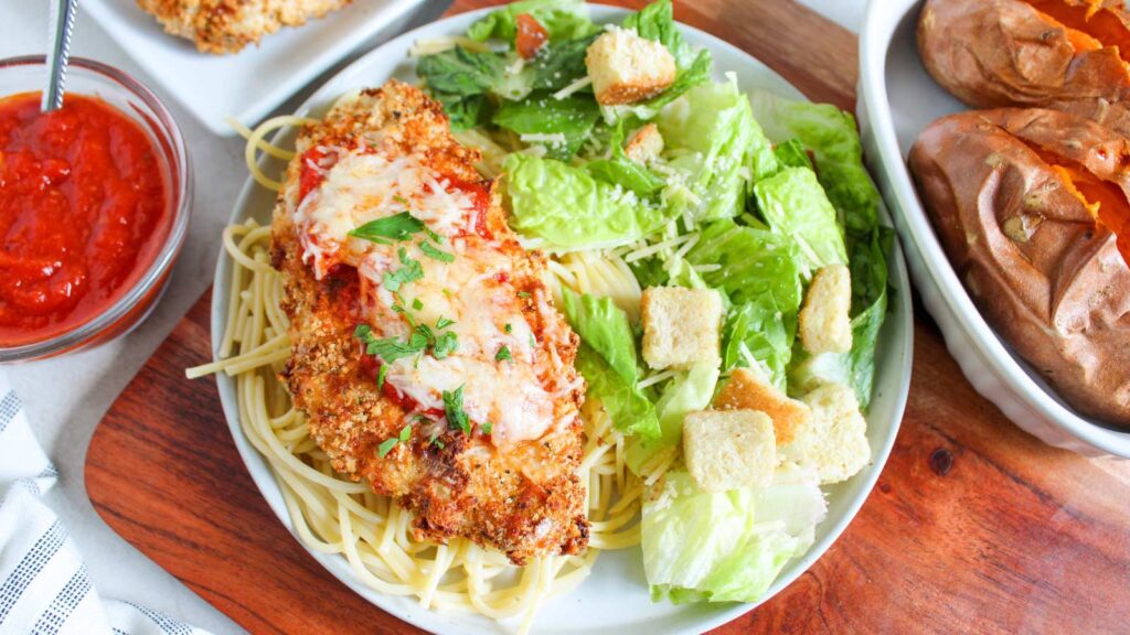 A white plate holds a breast of Air Fryer Chicken Parmesan on a bed of spaghetti noodles with a green salad next to it.