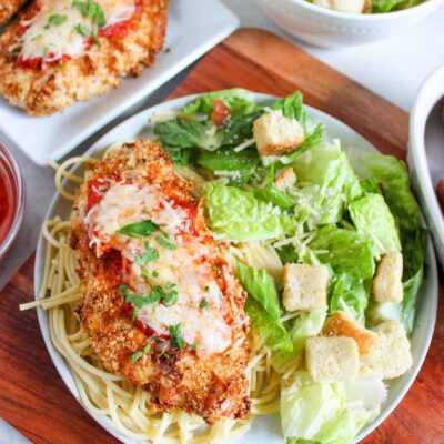 Overhead focus of a plate holding a breast of Air Fryer Chicken Parmesan on a bed of spaghetti noodles with a side of green salad.
