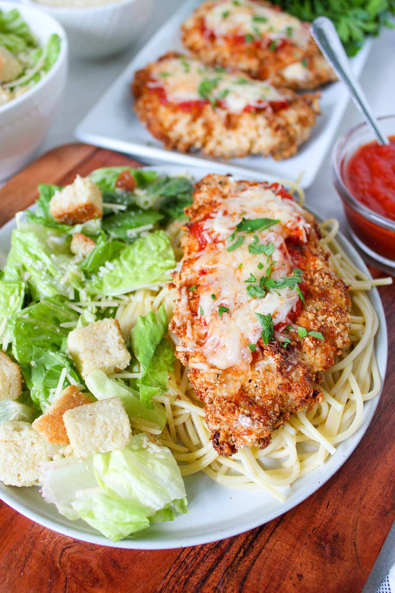 One breast of Air Fryer Chicken Parmesan laying on a bed of spaghetti noodles with a side of green salad.