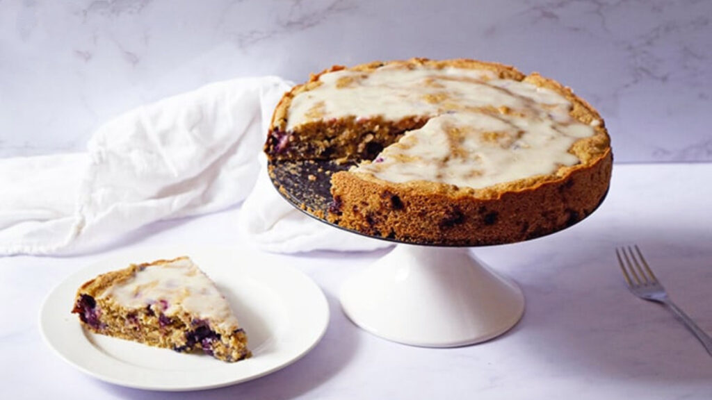 Blueberry Cake on a cake stand with a slice removed and place on a white plate sitting next to it.