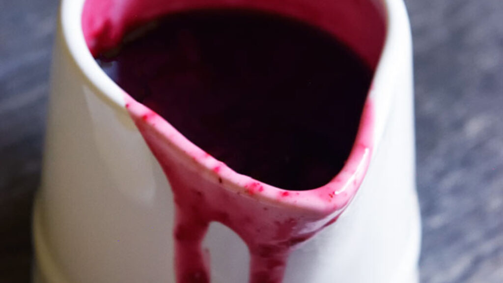 A single white pitcher filled with Blueberry Syrup. A bit of syrup has spilled over the edge of the pitcher and is dripping down the front.