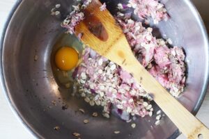 Mixed meatball mixture with an egg added to a mixing bowl.