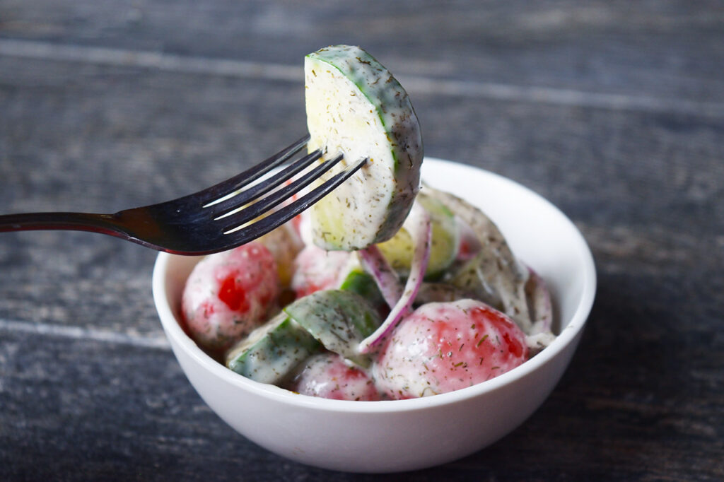 Side view of a fork lifting a cucumber from a white bowl full of cucumber salad.