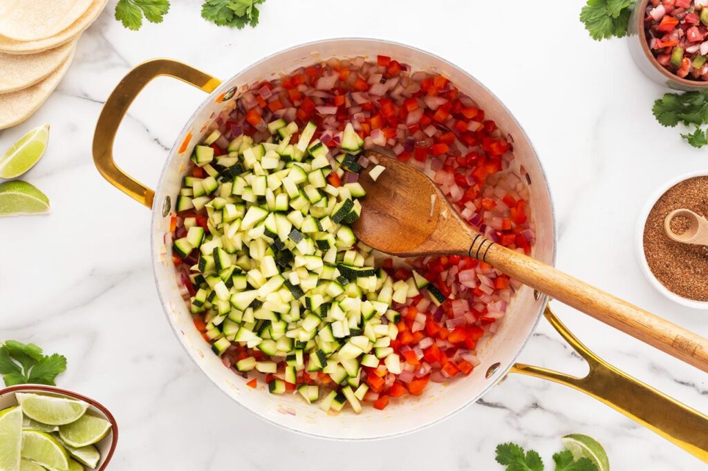 Adding zucchini to a skillet of sautéing veggies.