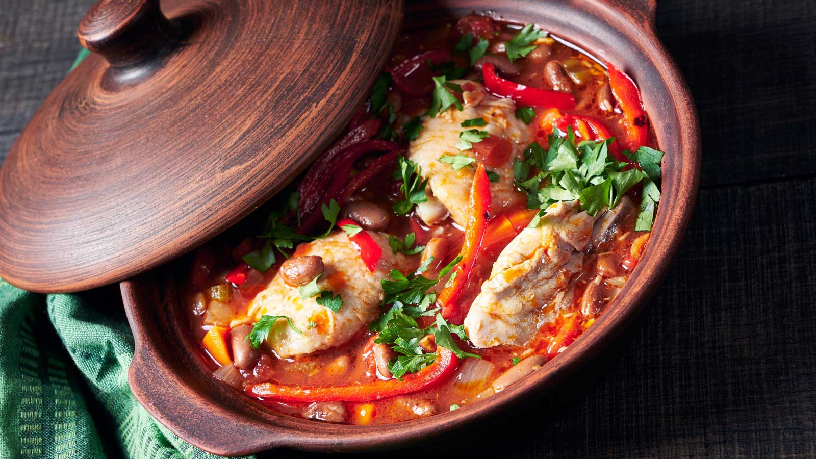 Healthy chicken stew of chicken breasts stewed in tomato sauce with vegetables and red kidney beans served on a rustic clay pot on a dark wooden background, top view, horizontal orientation.