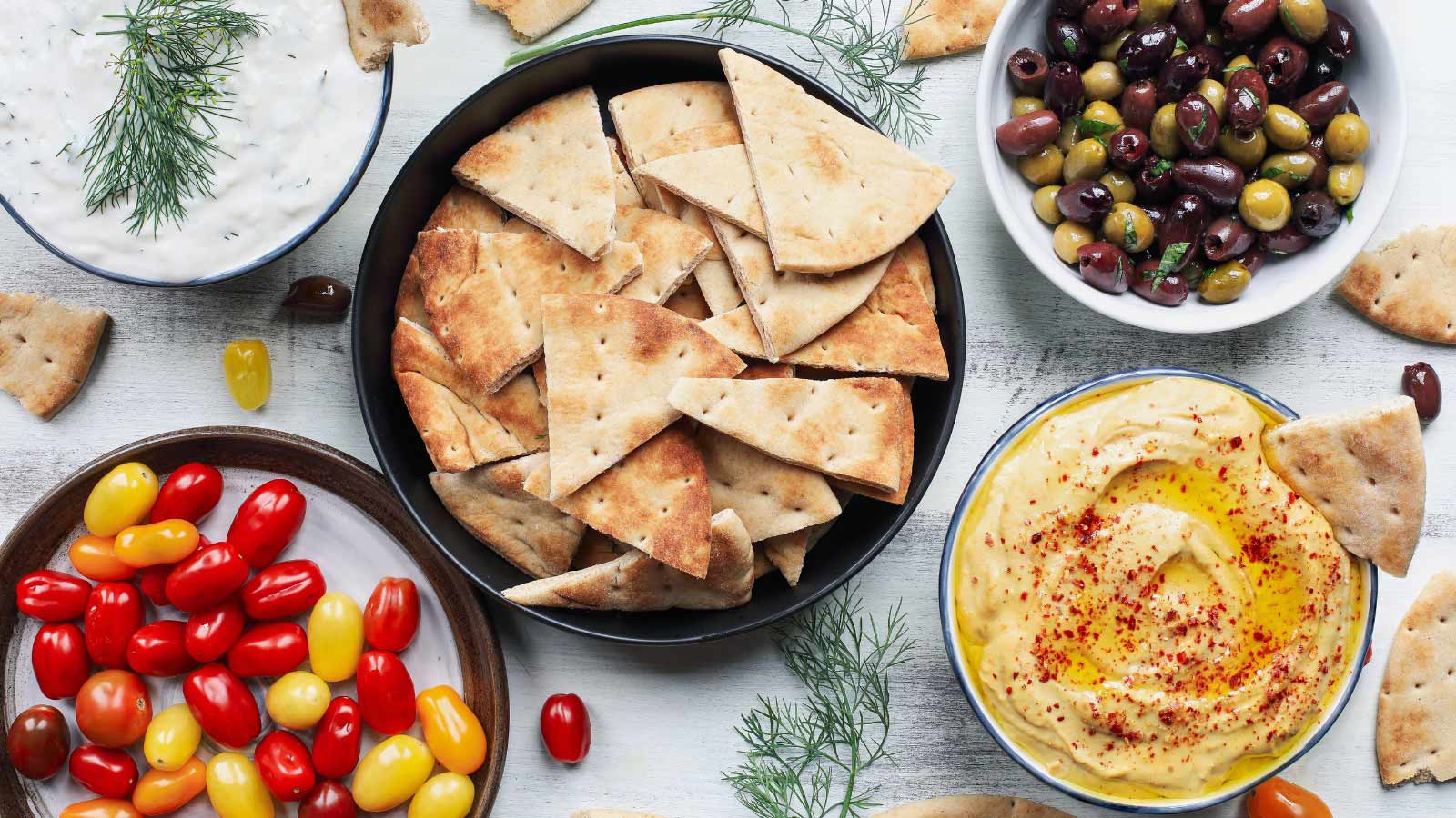 Mezze platter of pita bread surrounded by fresh tomatoes, olives, vegan tzatziki dip, and hummus over a white rustic table.