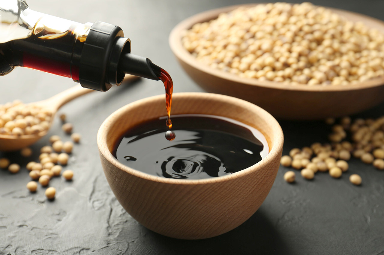 Pouring tasty soy sauce from bottle into bowl on black table, closeup.