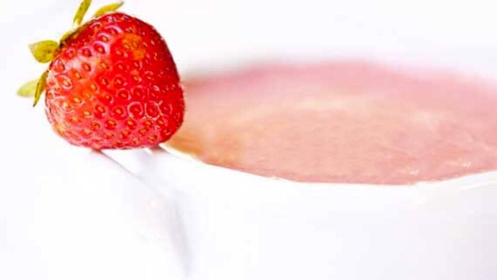 A white bowl holds a serving of strawberry soup. A fresh strawberry rests on the handle.