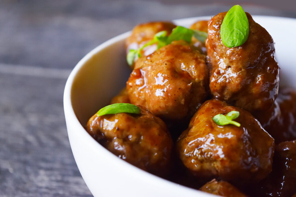 A side view of a white bowl filled with Sweet And Sour Meatballs.