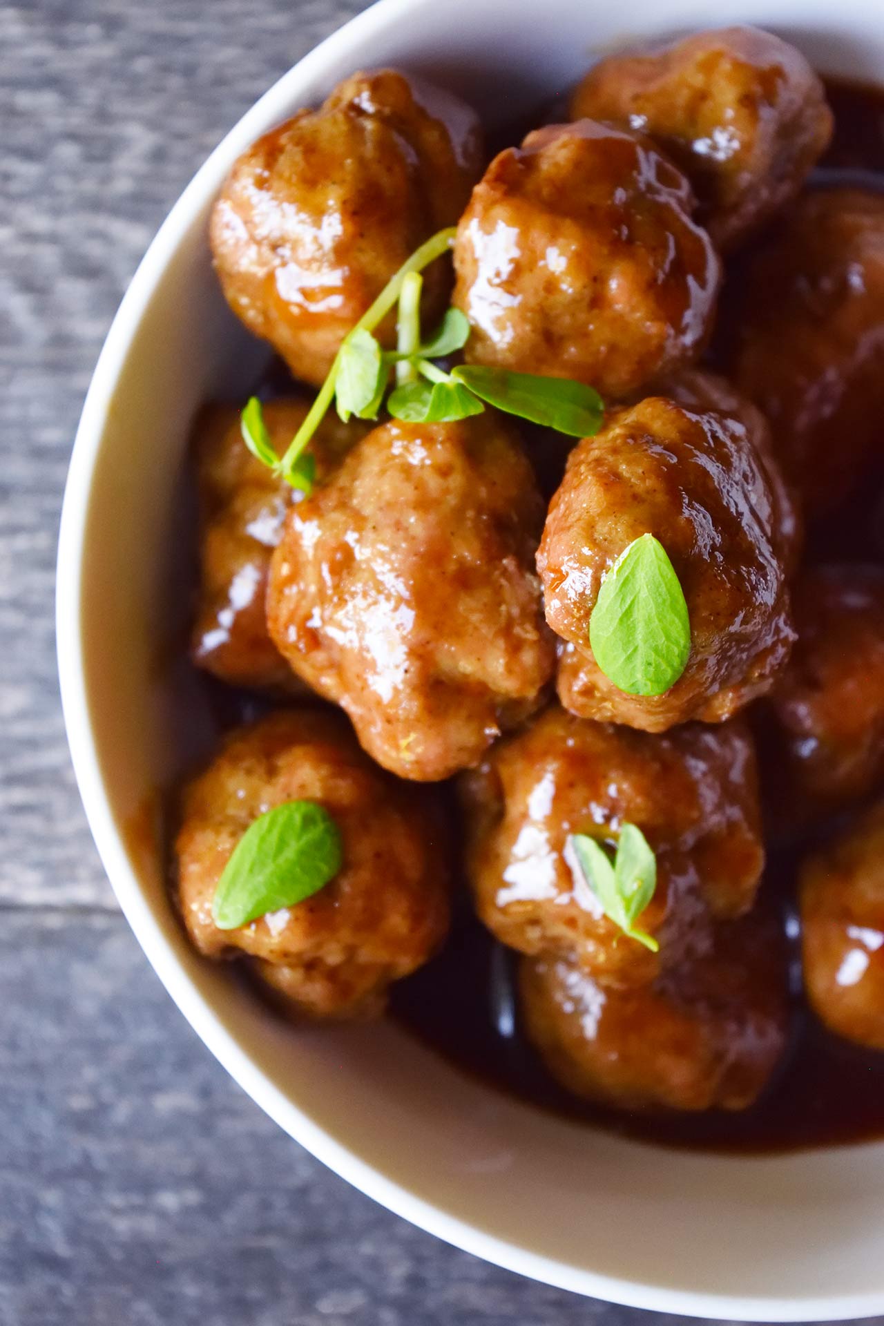 An overhead view of a white bowl filled with Sweet And Sour Meatballs and topped with a few leafy greens.