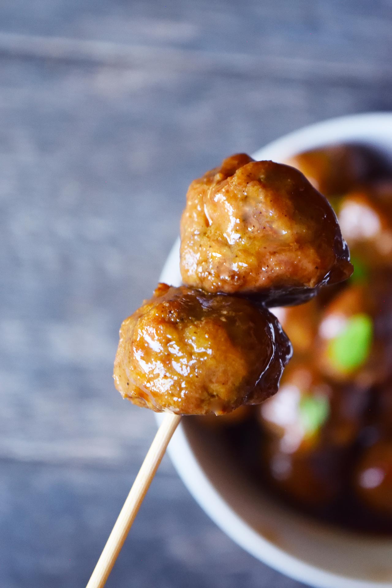 A wood skewer lifts two meatballs out of a white bowl full of them.