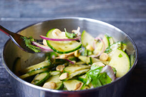 Side view of a fork lifting a bite of Thai cucumber salad from a metal bowl. Overhead focus.