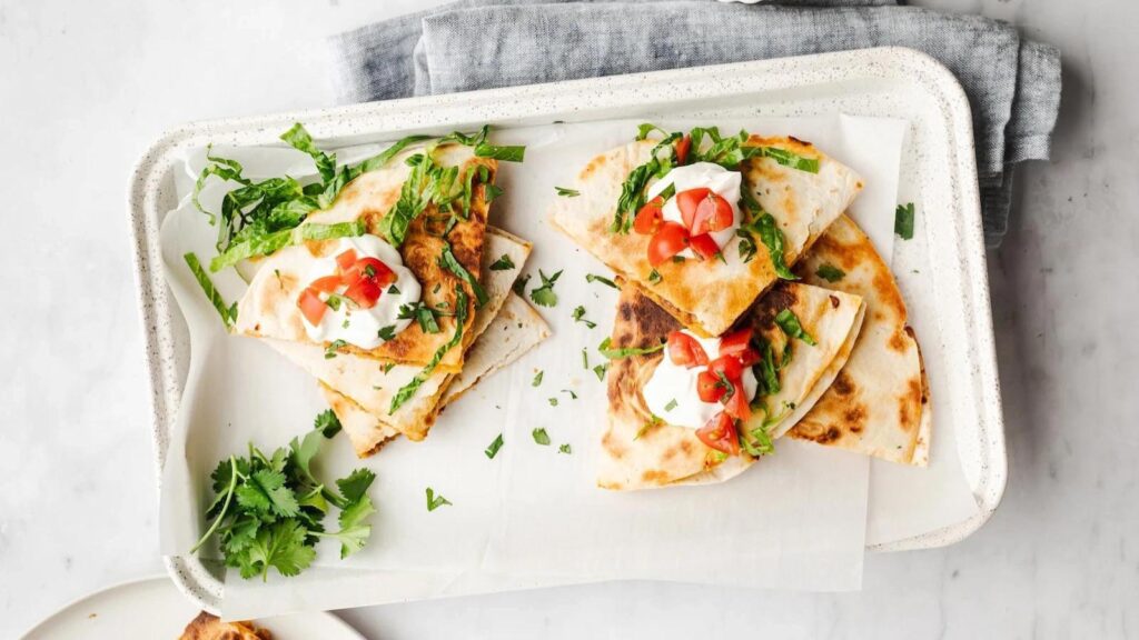 Overhead focus of cut beef quesadillas stacked on a platter with sour cream and tomatoes for toppings.