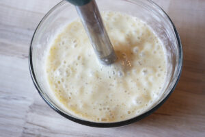 A stick blender blending bananas in a glass mixing bowl.
