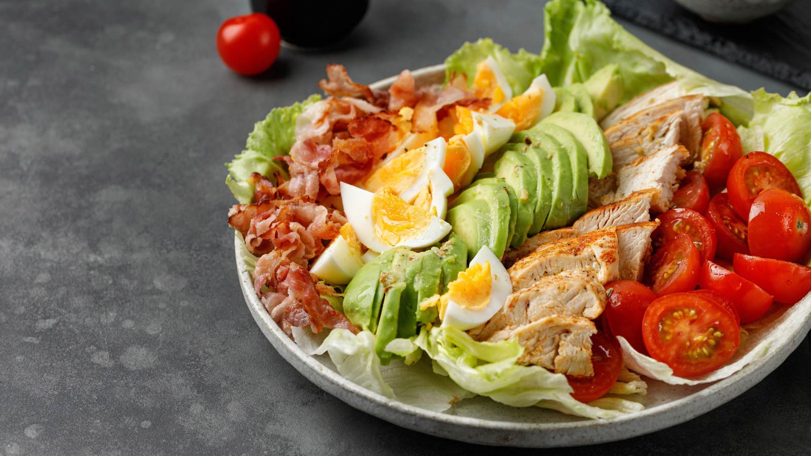 Cobb salad with avocado, bacon, tomato, grilled chicken, eggs on dark grey background.