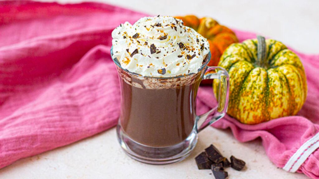 Whipped cream added to the top of a glass mug filled with pumpkin hot chocolate.