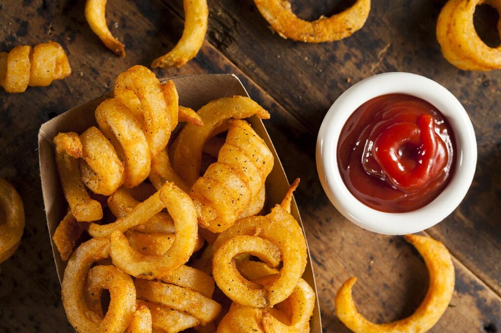 Curly fries ready in a carton with a small ramekin of ketchup next to it on a wood surface.