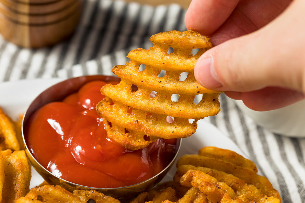 A hand dipping a waffle fry into some ketchup.