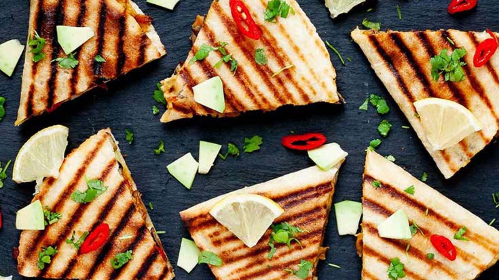 Cut hummus quesadillas laid out on a slate board with avocado pieces and lemon slices for garnish.