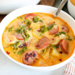 A white bowl with a spoon, filled with Sausage, Kale And Potato Soup.