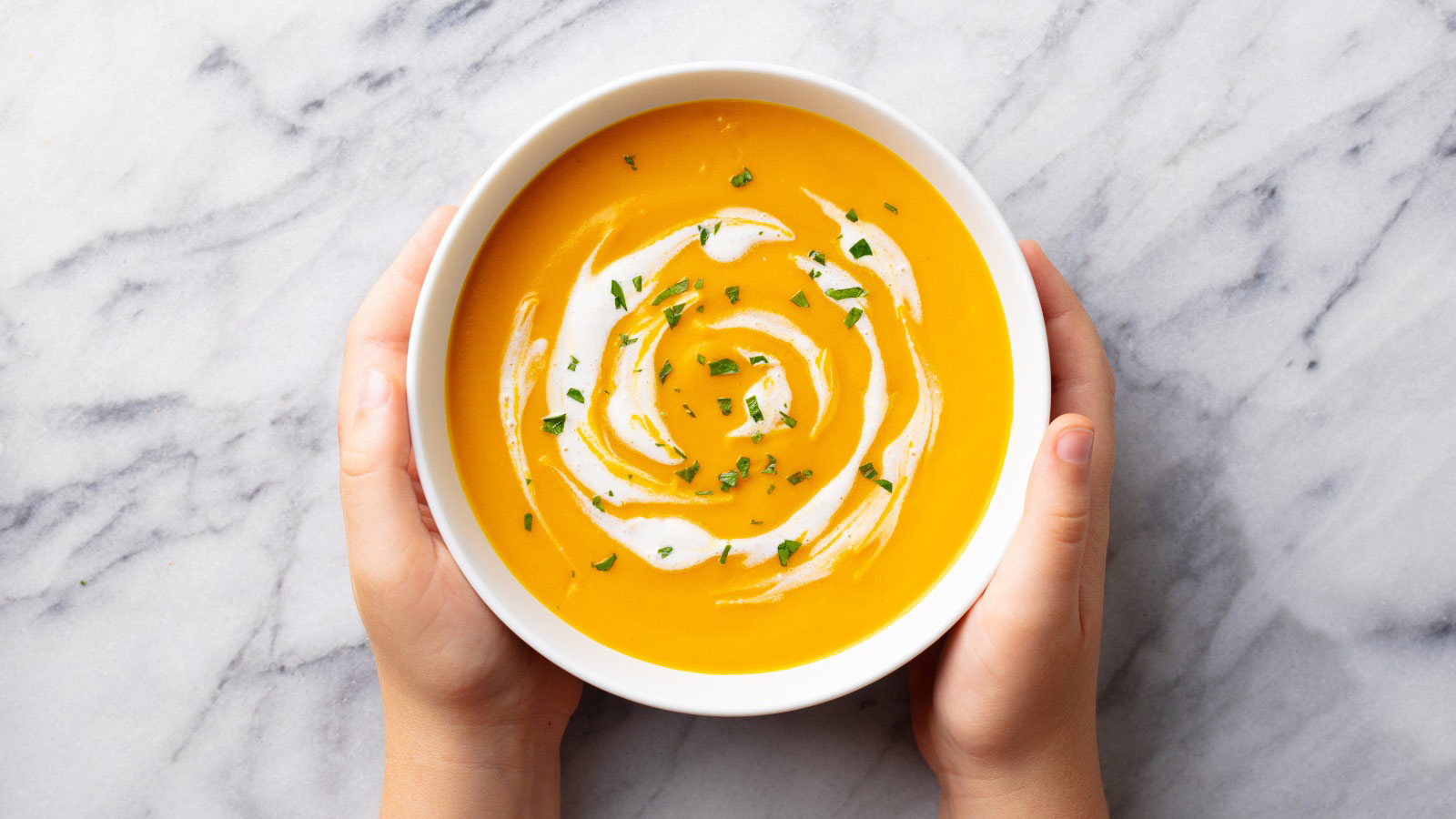 Kids hands holding a bowl with pumpkin soup. Marble background. Top view.