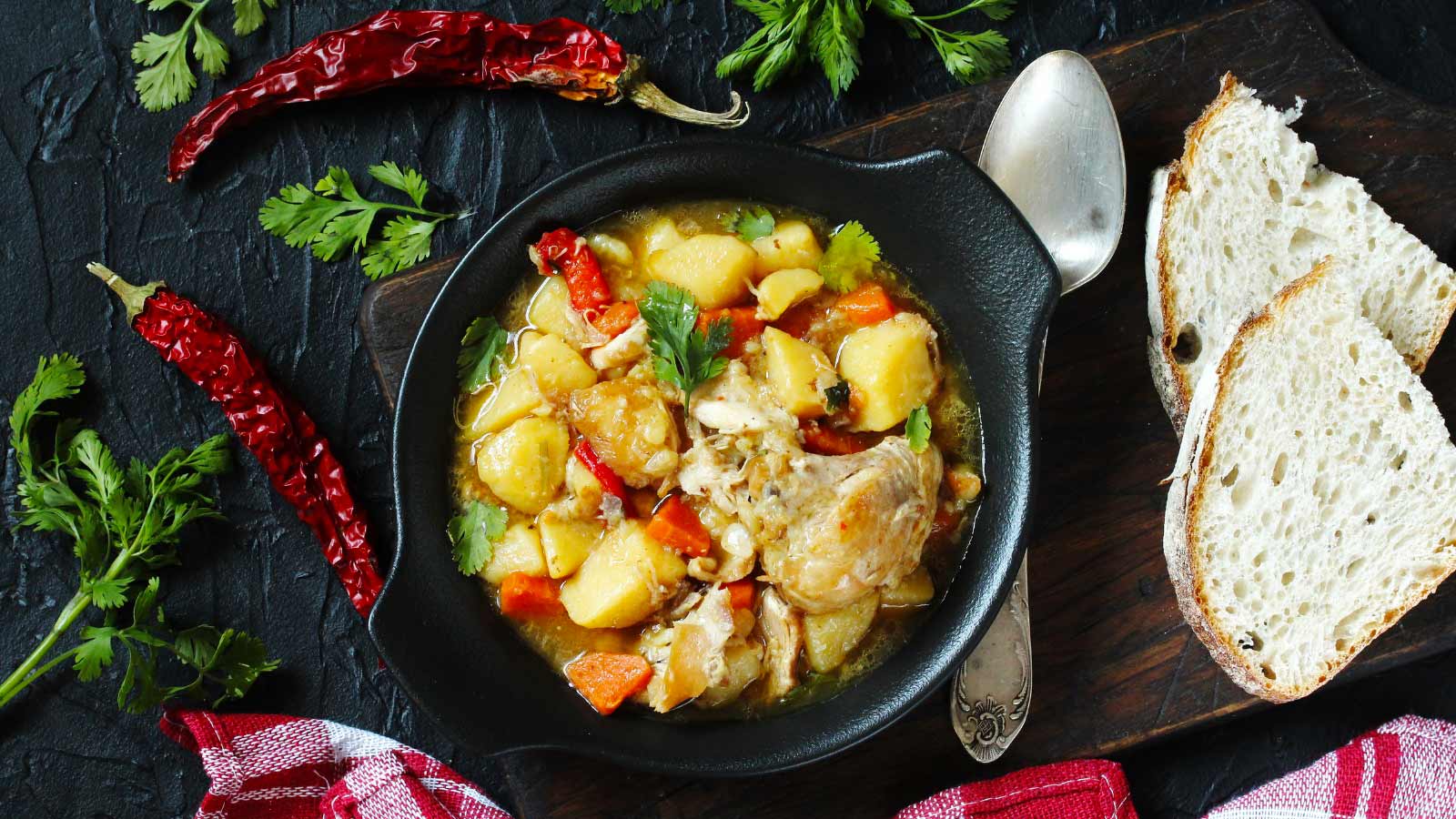 Chicken stew with potatoes and vegetables. in a black crock on a dark background surrounded by spices. Top view.
