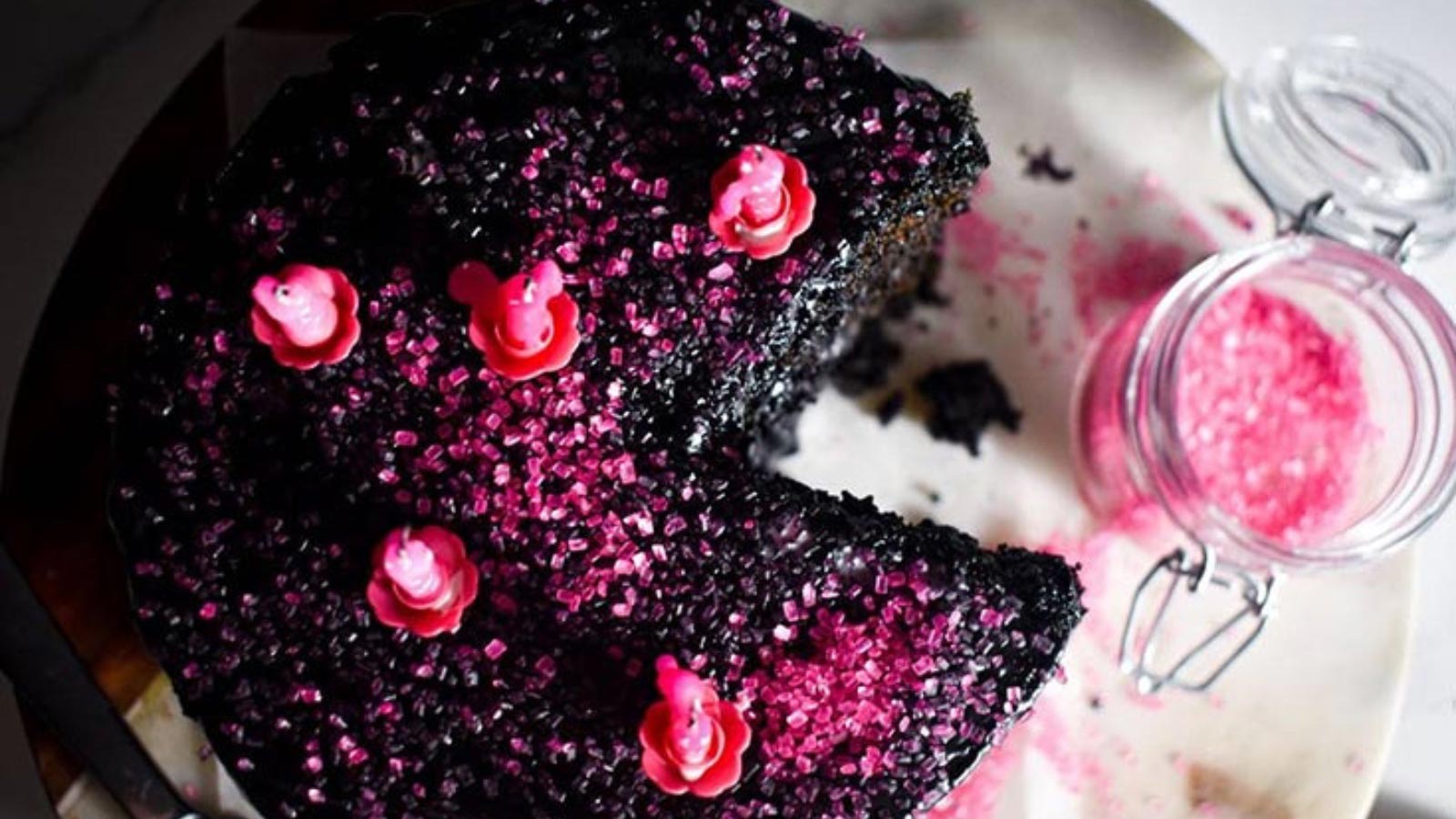 An overhead view of a chocolate cake with pink sugar and candles on top of black frosting.