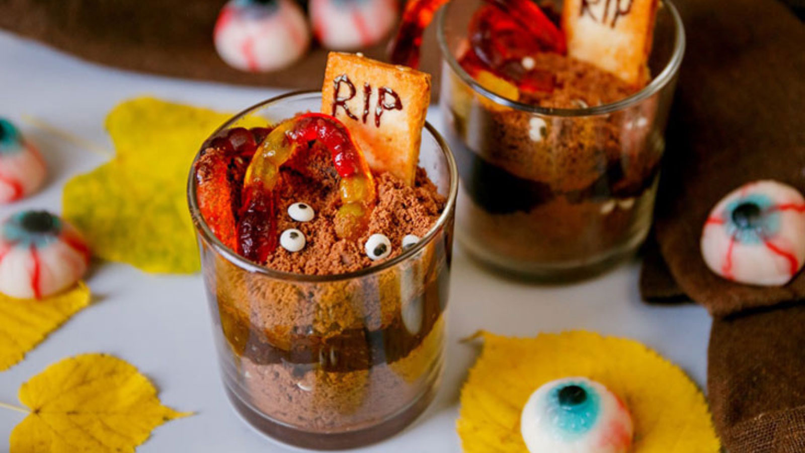 Two glass cups with chocolate jello, cookie crumbs, candy eyes and worms on top.