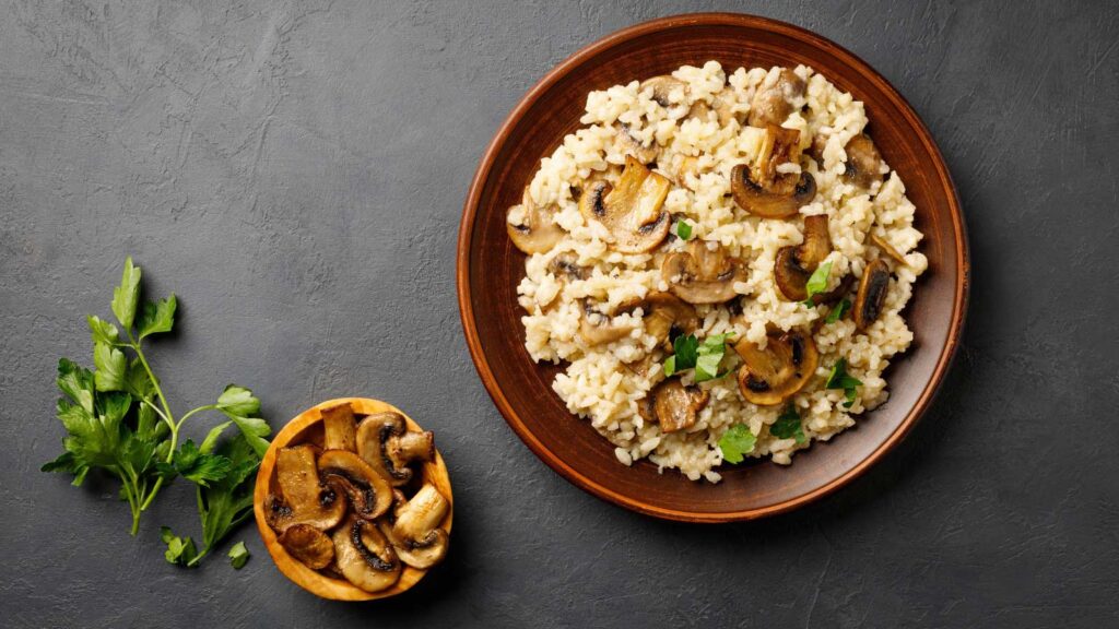 Brown rice and mushroom casserole served in a brown bowl with a smaller bowl of mushrooms to the side. Black stone backround.