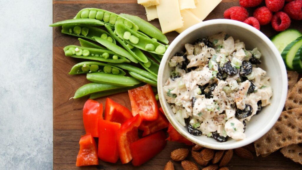 A white bowl filled with rotisserie chicken salad and surrounded by fresh veggies, crackers, cheese and raspberries.