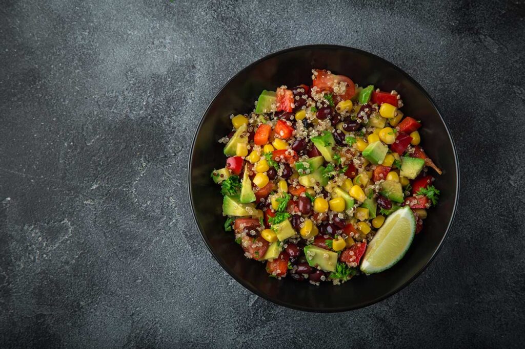 Avocado, quinoa, bean, corn and bell pepper dense bean salad in a black bowl on a slate surface.
