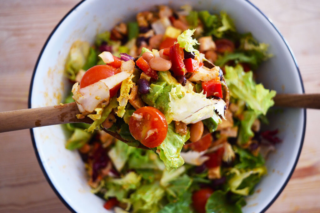 Overhead focus. White mixing bowl full of Mexican Dense Bean Salad. A wood spoon lifts up a spoonful.