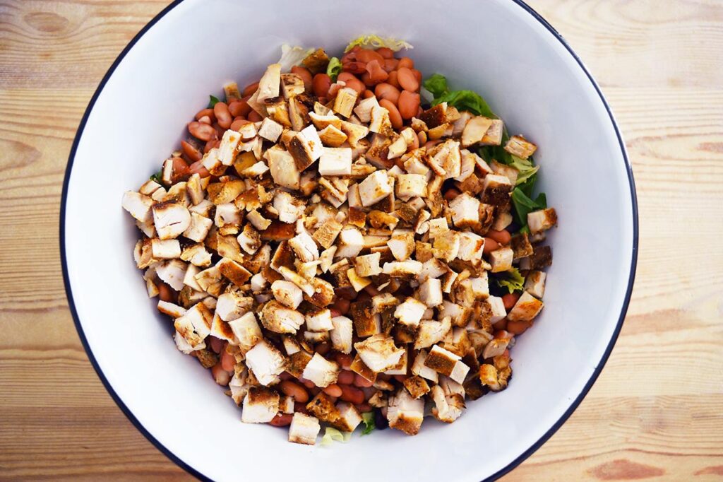 Chopped chicken breasts layered over beans and lettuce in a mixing bowl.